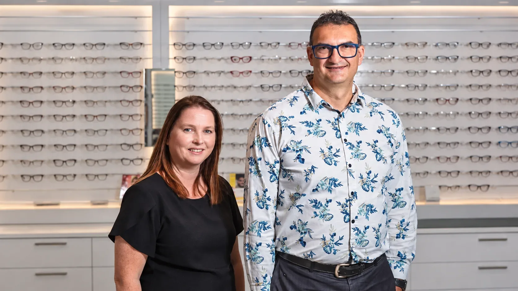 Two smiling team members in front of an eyewear display wall at Eyes by Design