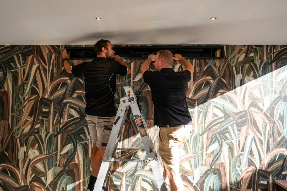 Two DPK installers fitting a ceiling air conditioning unit above a patterned feature wall