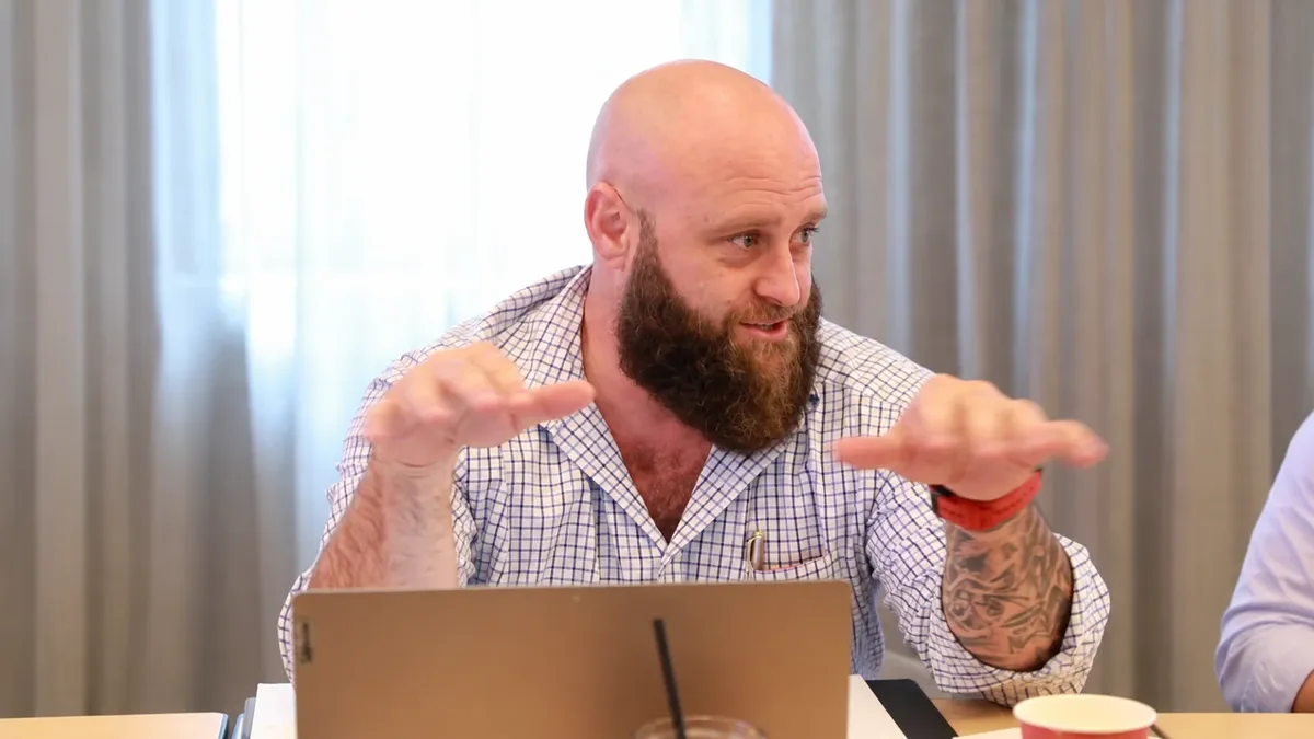 A man in a checked shirt talking at a workshop table, laptop open