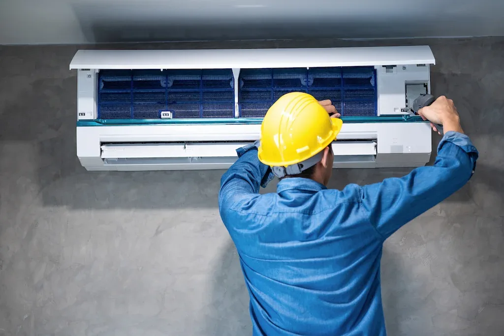 A technician in a yellow hard hat servicing a wall-mounted air conditioner